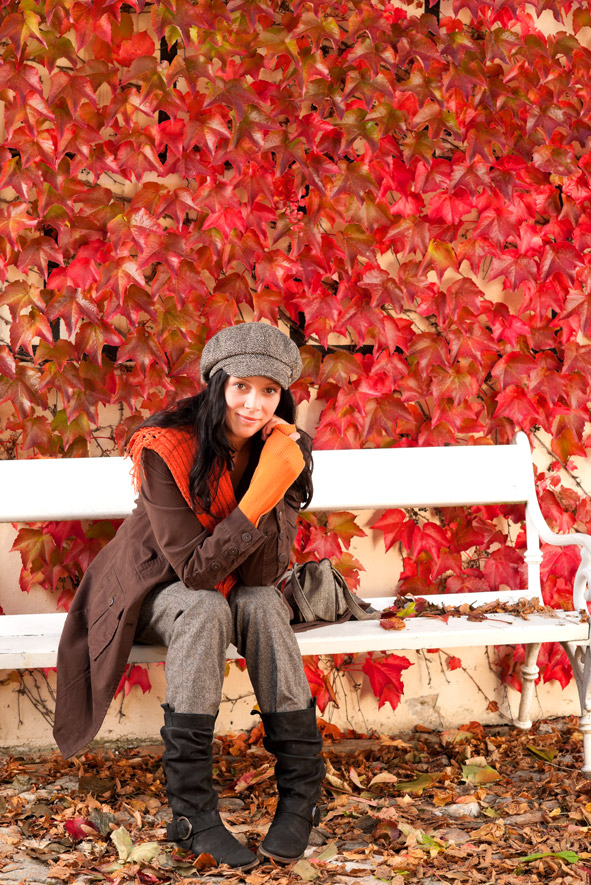 Autumn fashion young woman sitting on white bench in front of red colored wild wine leafs